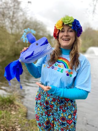 A person smiles at the camera. They wear bright blue clothing and a rainbow headband. They hold a handmade whale made from a recycled milk carton.