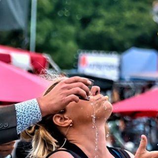 Two street artists perform in the middle of a festival. One performer stands behind the other pouring water into their mouth while they juggle with kitchen whisks.