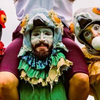 Three performers dress up as birds, they stand in a huddle. Each performer has a headpiece, one in red, one in yellow and one in green.