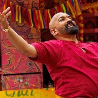 A performer stands with their arms open wide looking to the sky. They wear a vivid red t-shirt and stand infront of a bright yellow TukTuk.